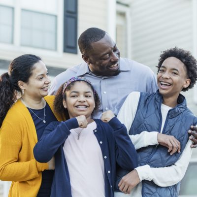 An interracial Hispanic and African-American family with two children standing on the driveway in front of their house. The father is African-American, mother is Hispanic, and the 14 year old teenage boy and his 11 year old sister are both mixed race. Both children are wearing braces. The girl is looking at the camera.